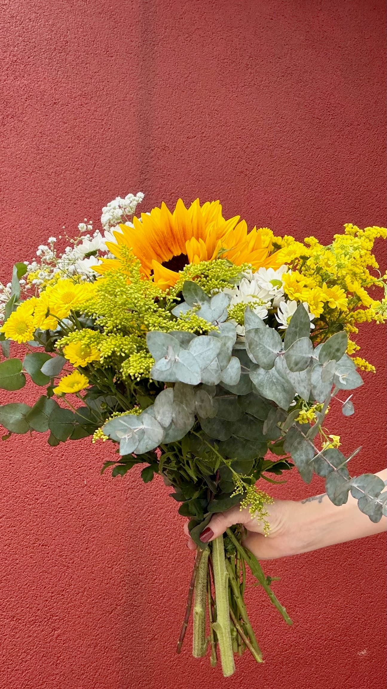 Buquê autoral em tons de amarelo, branco e verde diante de uma parede vermelha